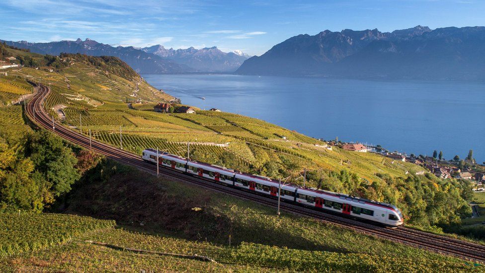 A Swiss train passes a lake through the countryside, with mountains in the background.
