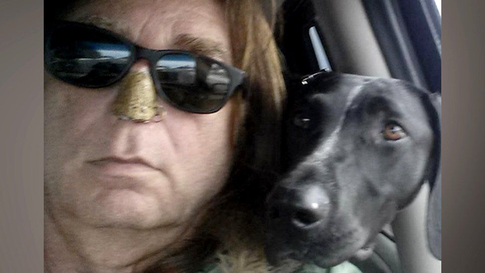 James Jacobson, pictured wearing sunglasses and with his trademark snakeskin nose cover, sitting in a car with a black dog