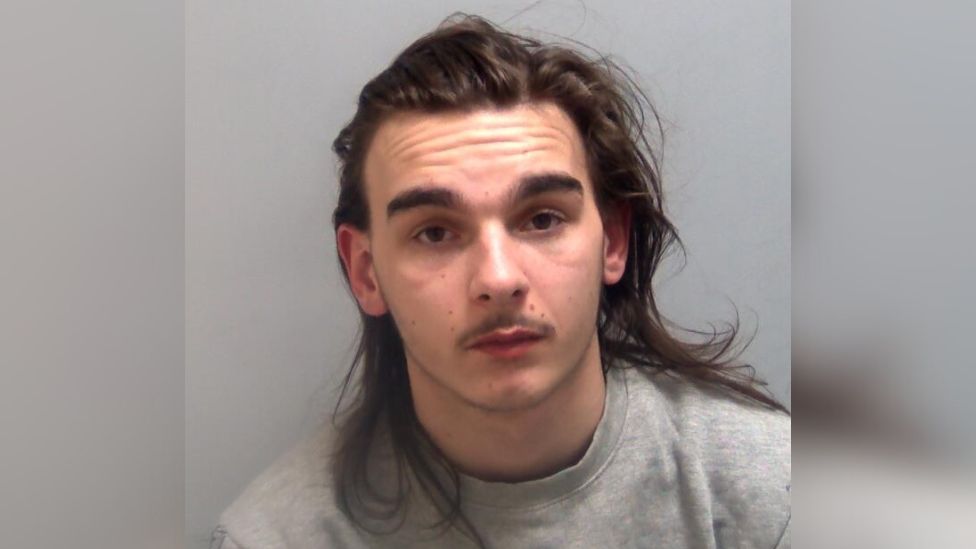 A custody image of Callum Taylor, who has long, dark hair. He is wearing a grey sweatshirt and looking expressionless at the camera.