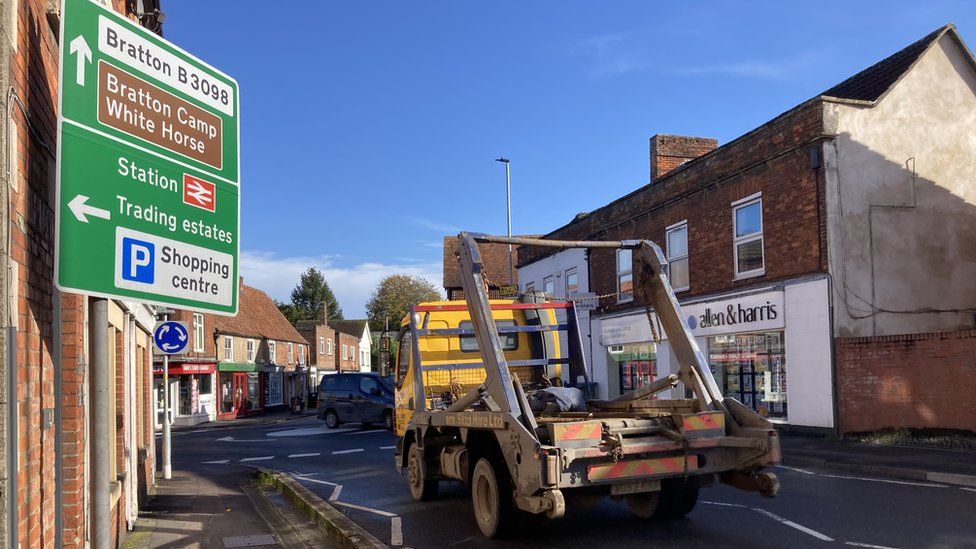 A truck that carries skips approaches a mini-roundabout in Westbury along the A350 with a road sign on the left.