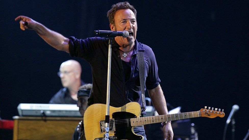 Bruce Springsteen sings into a microphone while holding a guitar