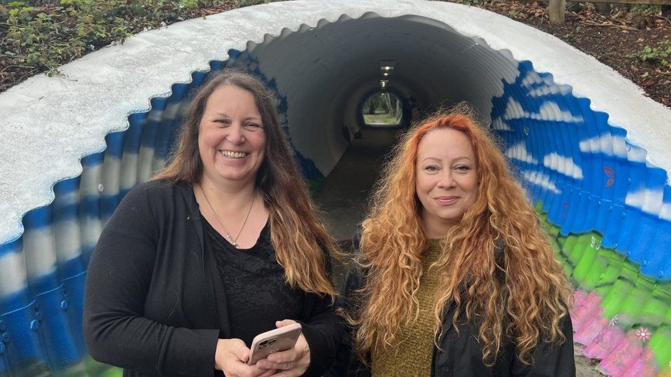 Two women, one with brown hair and one with orange, stood in front of a blue painted tunnel