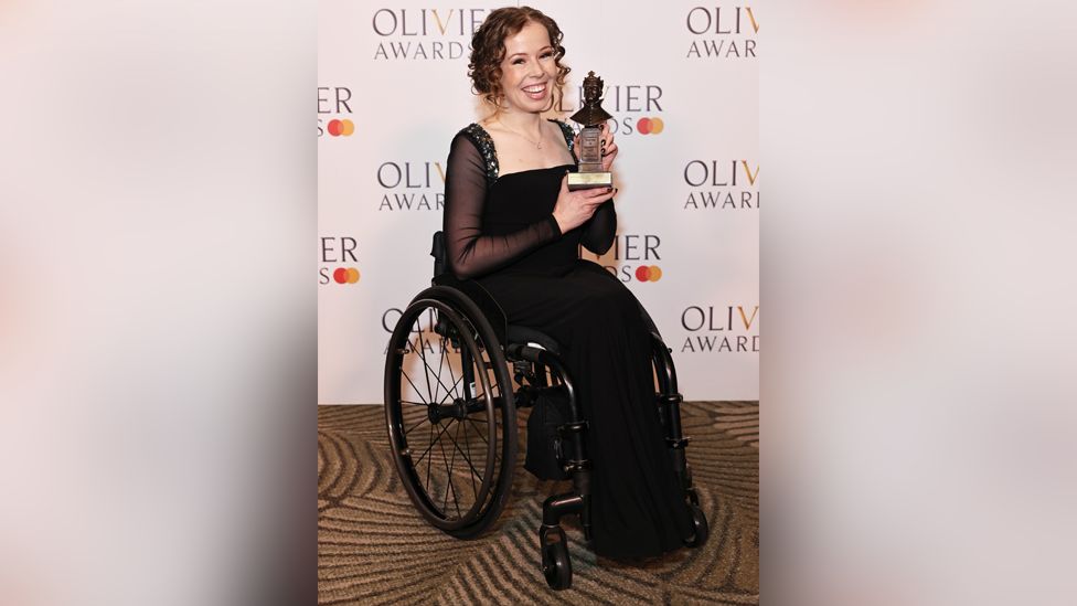 Amy Trigg wearing a black dress while holding her Olivier award next to her face and smiling at photographers