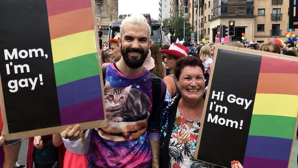 A mother and son hold signs. One says 'Mom, I'm gay' and the other says, 'Hi Gay, I'm Mom'.
