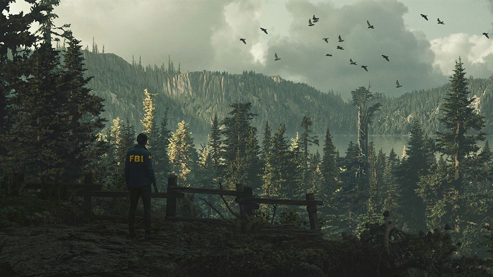 A woman in a blue jacket with FBI in yellow letters stands in a forested area looking out over the landscape below. It is densely populated with pine trees of varying heights, that totally cover the surrounding hills and valleys. The surface of a large lake can just be made out inbetween the tops of the trees in the distance. A grey, cloudy sky above creates a foreboding atmosphere and a flock of birds is seen flying through the frame.