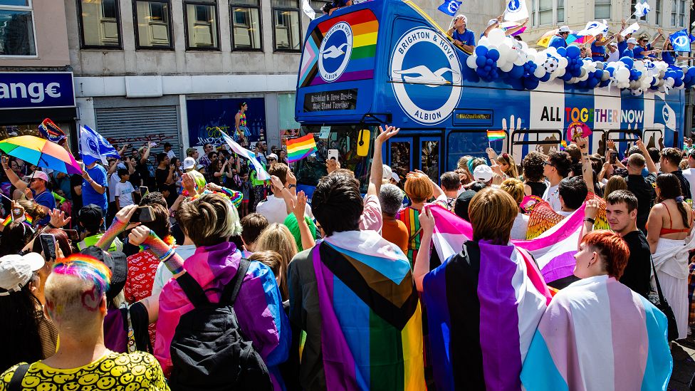 A Brighton & Hove Albion FC bus passes revellers draped with Pride flags during the 30th anniversary Brighton & Hove Pride LGBTQ+ Community Parade on 6th August 2022 in Brighton