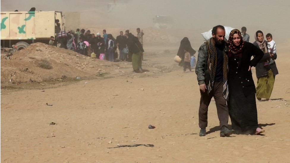 An Iraqi man and woman, who look to be struggling, walk amid dust and sand as they flee their homes in west Mosul. Dozens of people, including children are seen behind them.