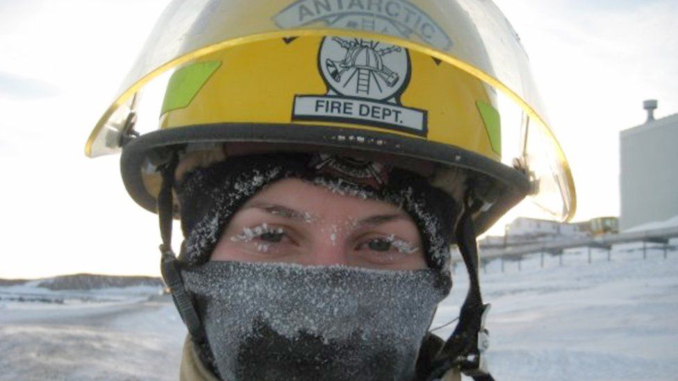 A woman in a yellow helmet with 'Antarctic Fire Dept.' written on it. She is wearing a scarf over her nose, covered in ice.