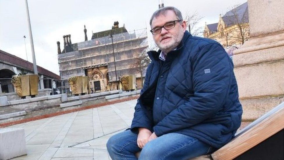 Man with short hair, beard and glasses sits in a public square with building work in the background