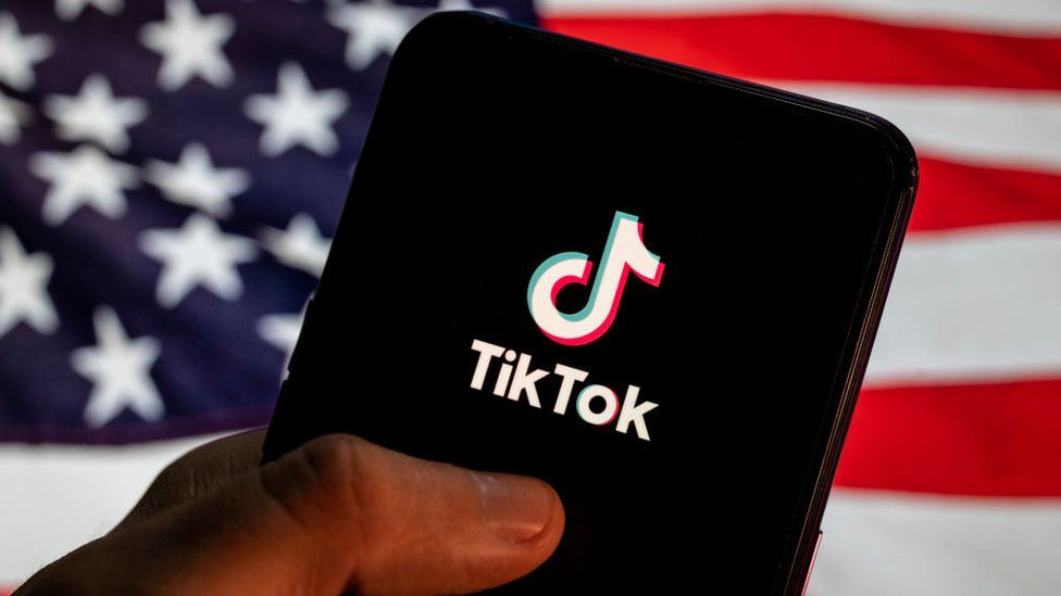 The TikTok logo is seen on a mobile device with the US flag in the background.