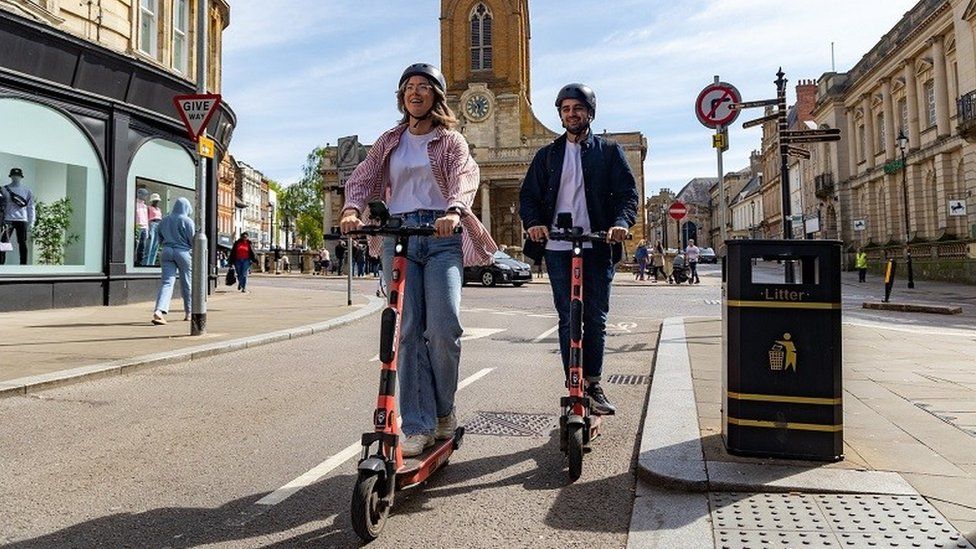 A man and a woman, wearing helmets, ride scooters up Abington Street in Northampton, from the Market Square