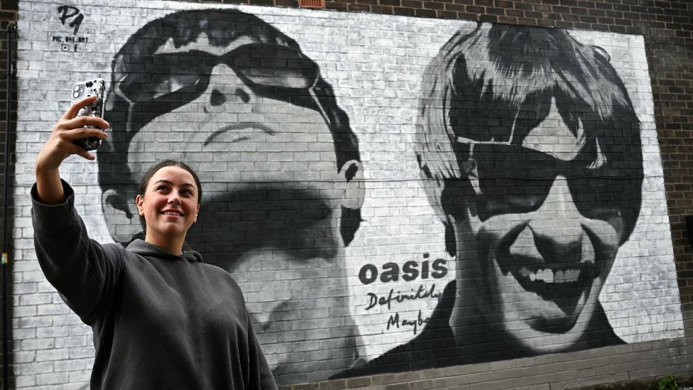 Fan Emily McShane, takes a photograph of a new street artwork depicting Oasis' Liam and Noel Gallagher, created by Manchester-based street artist Pic.One.Art. on the side of the Sifters Record store in Burnage, a suburb of Manchester, northern England on August 27, 2024