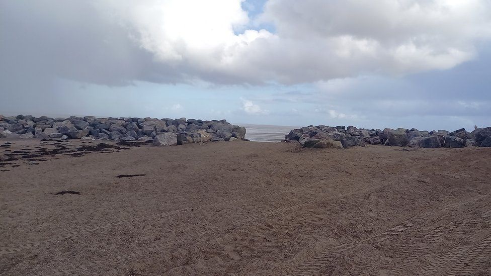 A row of grey boulders lined up along the shore on a sandy beach with a gap in the middle showing the sea