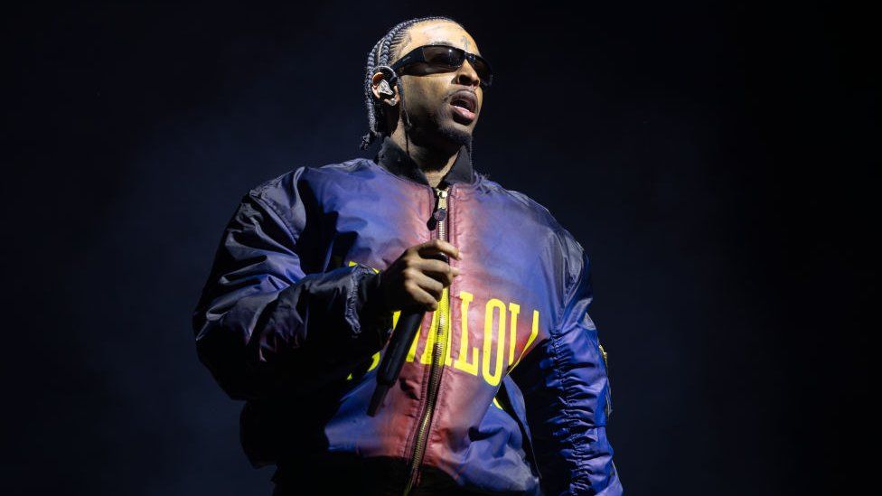 21 Savage performs his first ever London headline show at The O2 Arena on November 30, 2023 in London. The rapper has braided hair and wears sunglasses and a blue and red bomber jacket. He holds a microphone and is pictured on stage, which is dark behind him.