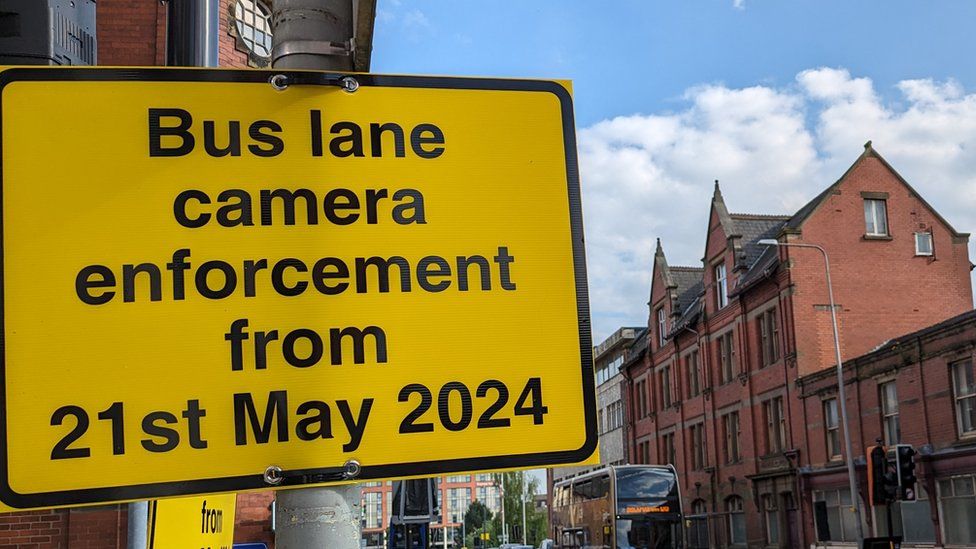 A yellow sign reads "Bus lane camera enforcement from 21st May 2024" on Corporation St in Preston.