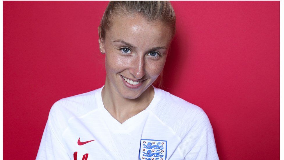 Leah Williamson wearing a white England shirt while standing in front of a red background