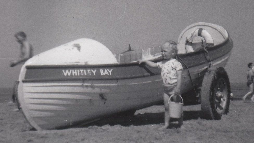 Black and white picture of a young boy with his hand on a boat on a beach