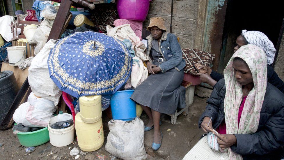Three women sit with a pile of buckets, bedposts and an umbrella outside a doorway