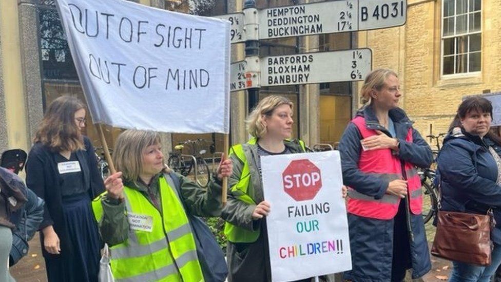 Five protesters standing outside a school with signs saying "stop failing our children" and "out of sight out of mind"