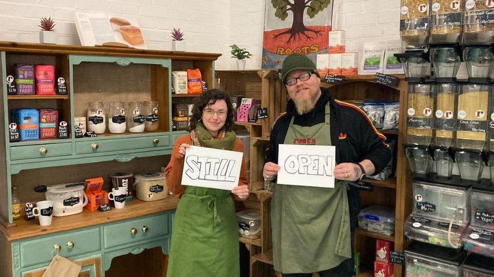 Two staff members at Roots refill standing in the shop holding signs that say "Still" and "open"