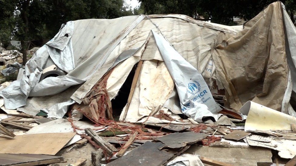 A dirty, damaged white tent with the UNHCR logo visible to the side. The surrounding area is littered with debris.