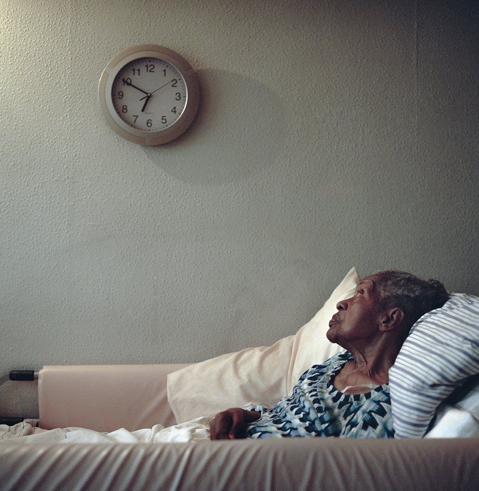An elderly lady lying in bed looking at a wall clock