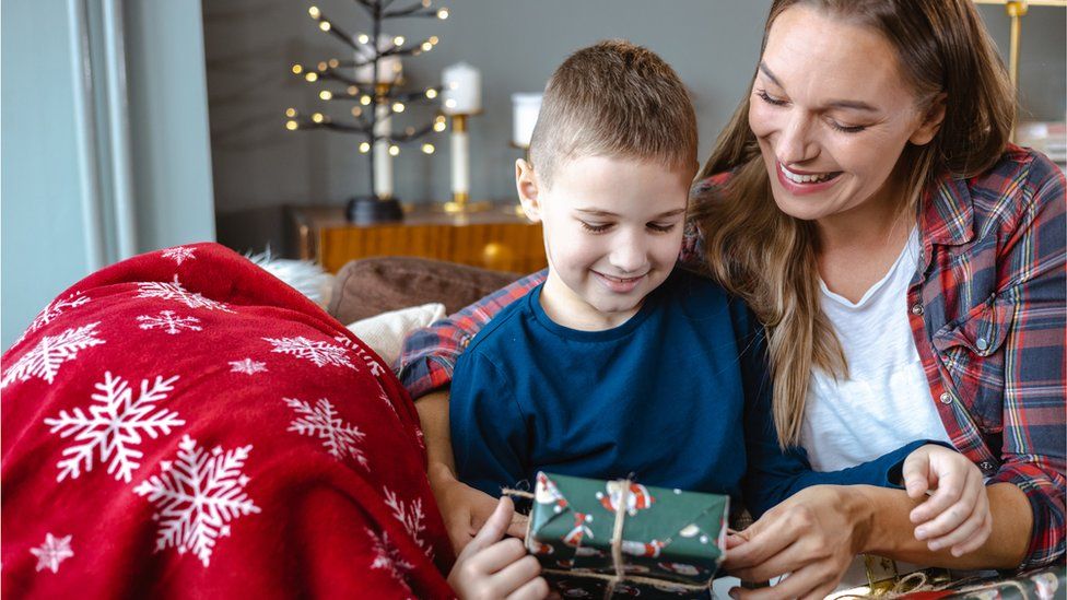 A woman is sitting on a living room sofa with her young Caucasian son and giving him Christmas presents.
