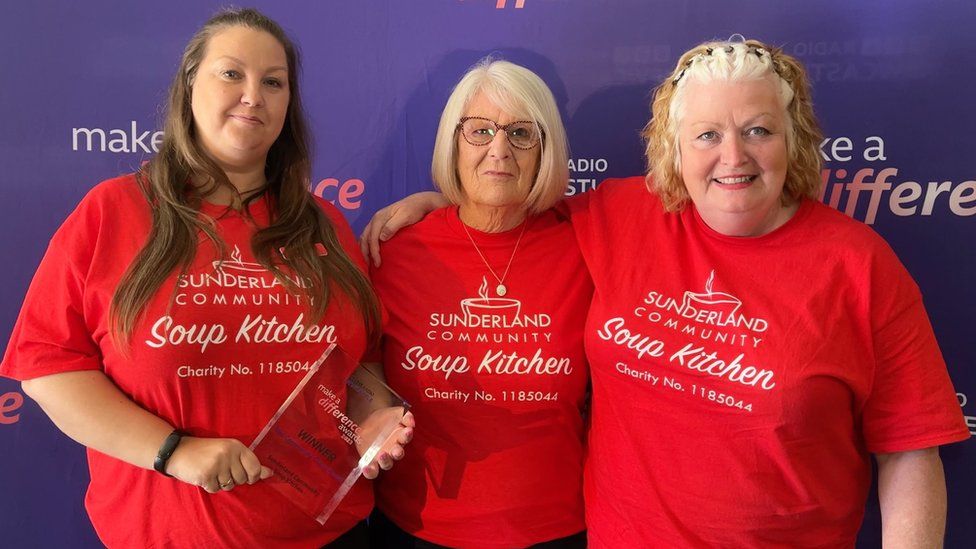 Three women in red t-shirts hold a glass award