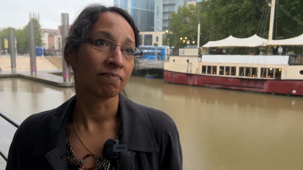 Jaya Chakrabarti with tied back dark hair looking off camera with a tight mouth smile wearing glasses standing in front of the Bristol harbour