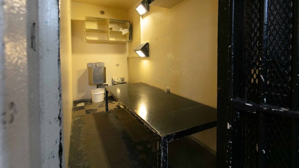 A photo of one of the cells, with a black table, a few lights, shelves, and a toilet