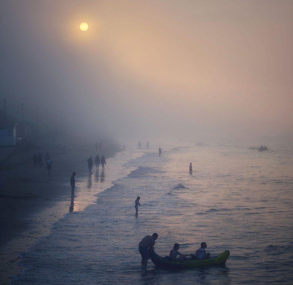 People playing in the sea on a misty day with the sun a disc as seen through cloud