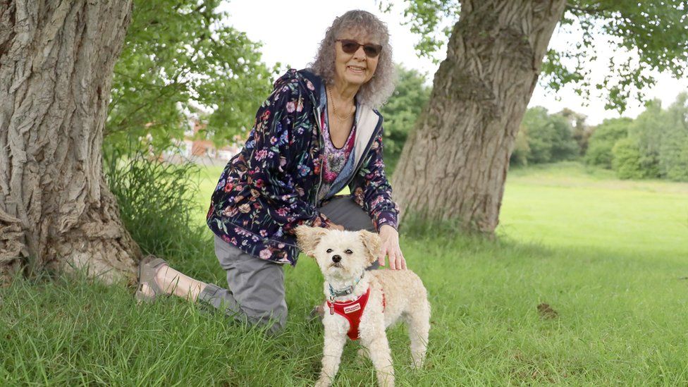 Pip in the park petting a small white poodle under some trees.