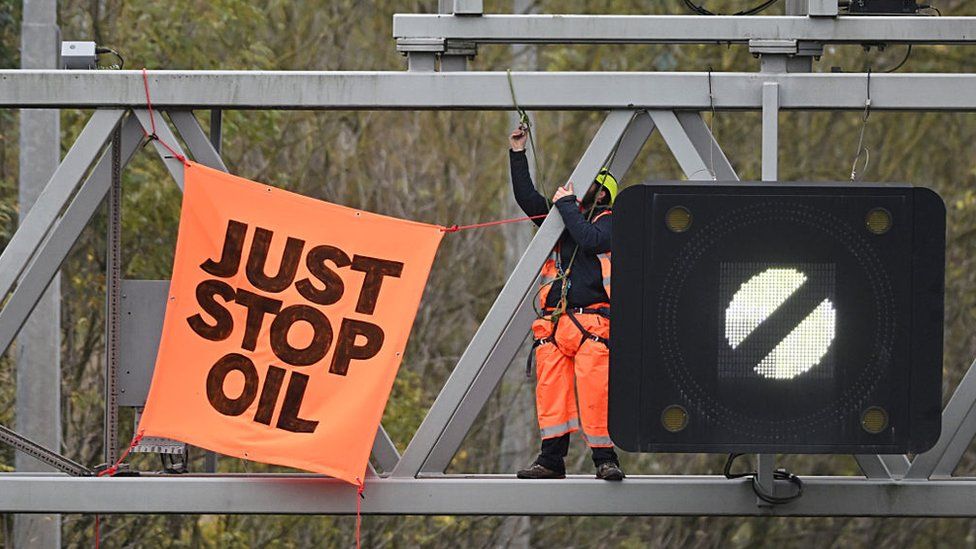 An activist puts up a banner reading "Just Stop Oil" atop an electronic traffic sign along M25 on 10 November