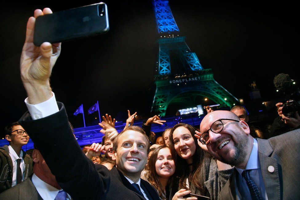 French president Emmanuel Macron uses his iPhone to take a selfie with the crowd in front of the Eiffel Tower