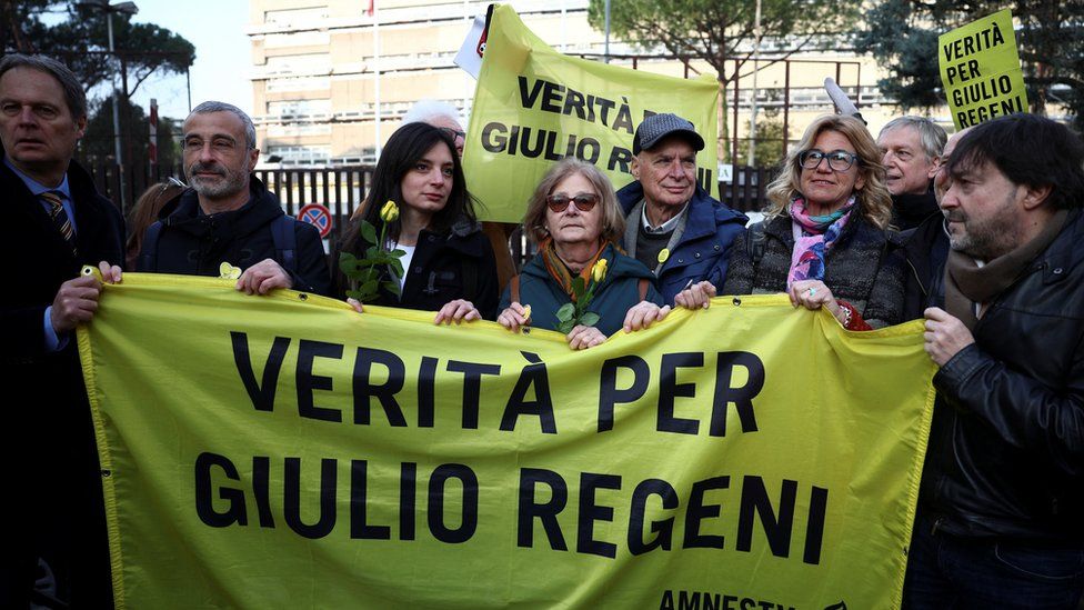 Mr Regeni's family and others hold yellow roses and a banner outside court. The banner reads: "Truth for Giulio Regeni".