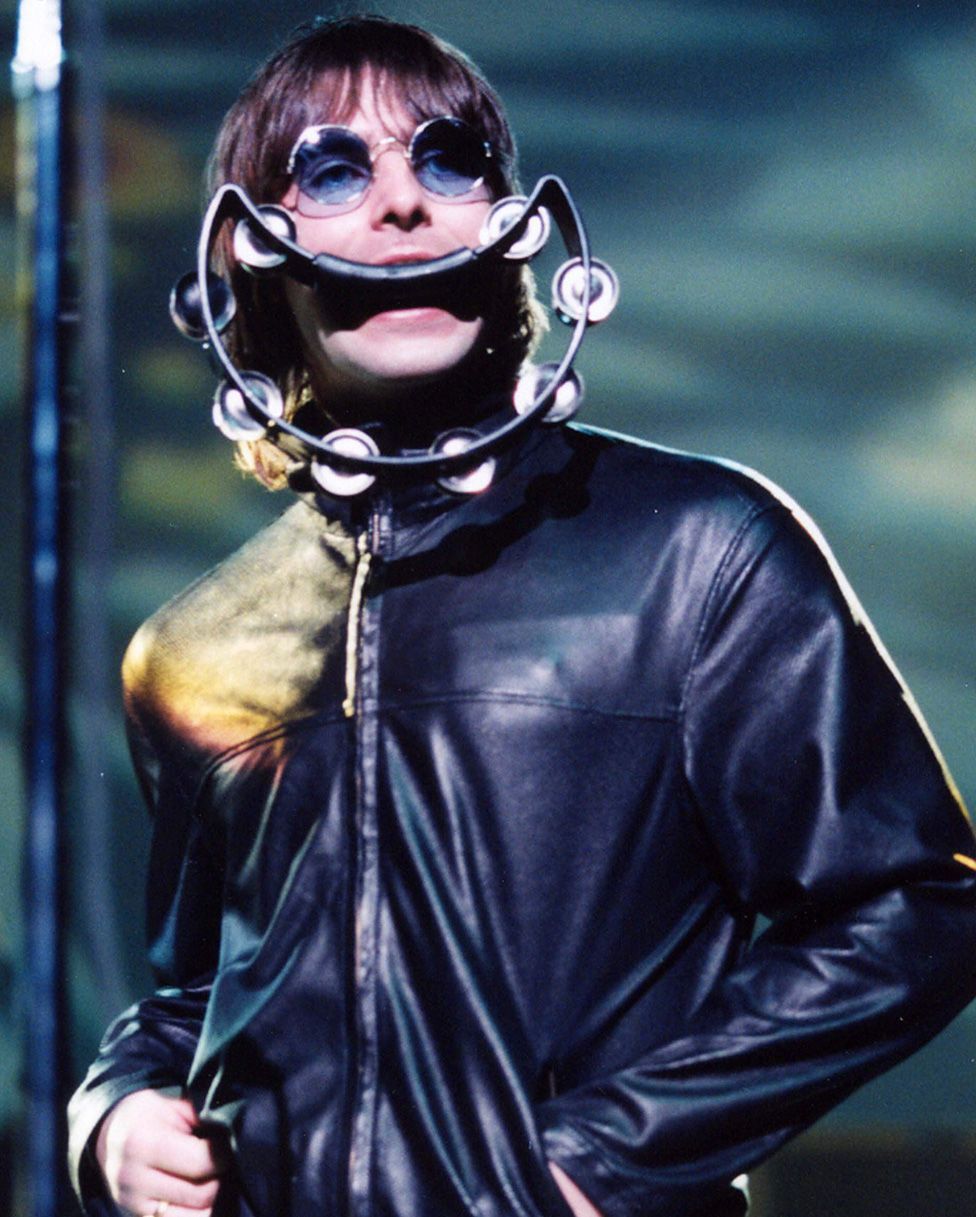 Liam Gallagher on stage in 2000 with a large crescent-shaped tambourine in his mouth, looking like a giant smile