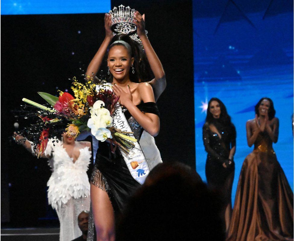 Miss SA Ndavi Nokeri smiling in a sash with flowers as she received her crown. There are women behind her clapping.