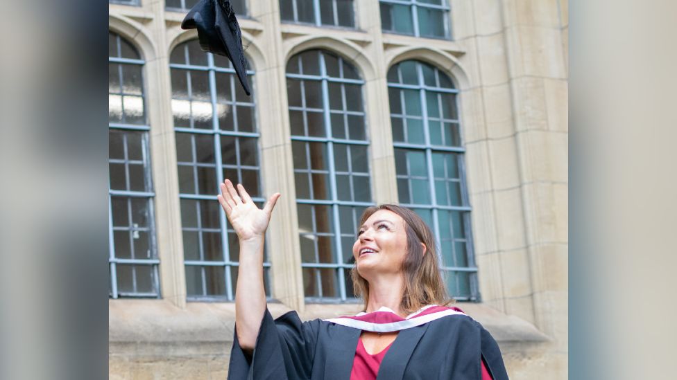 Dr Becs Bradford pictured at her university graduation. She is wearing a red dress with a black graduation gown over the top. She is looking up, with her hand up, after throwing her graduation cap in the air. 