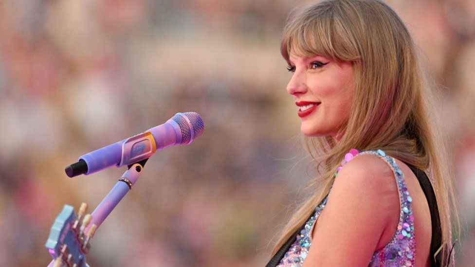 Taylor Swift on stage, her side profile is facing the camera and she's smiling with a microphone placed in front of her