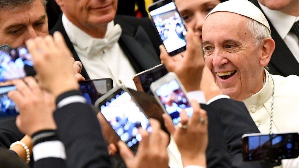 Pope Francis smiles as people take pictures on their mobile phones