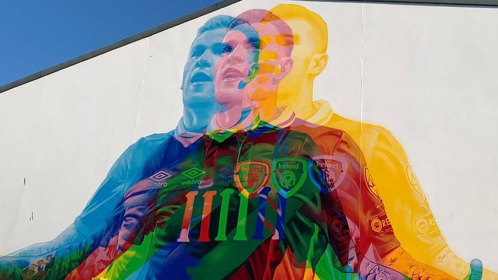 A mural on a white wall showing James McClean wearing an Ireland jersey. The mural is made up of three images of him in his kit - one is red, one is blue and one is yellow. His arms are out at his side and his mouth is slightly open in what could be a goal celebration. Where the pictures overlap the colours mix to create new ones and give the overall image a holographic, rainbow effect.