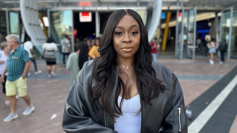 Rachel Otto, pictured outside Asake's concert at the O2 Arena in London. Rachel, a black woman in her 20s, has long black hair almost down to her middle. She wears a black leather bomber jacket over a low cut white top and wears a silver necklace with the letter 'R'.