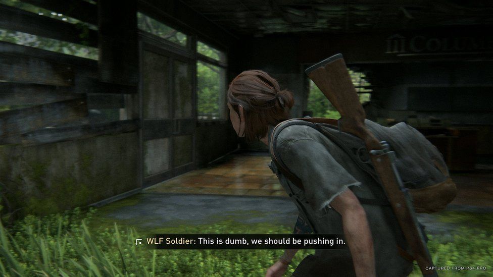 In this screenshot, the main character of the Last of Us Part 2 is seen sneaking through a run-down shack with a rifle on her back. Subtitles show a solider saying "this is dumb" - with an arrow on-screen indicating the sound is coming from the other side of a wall