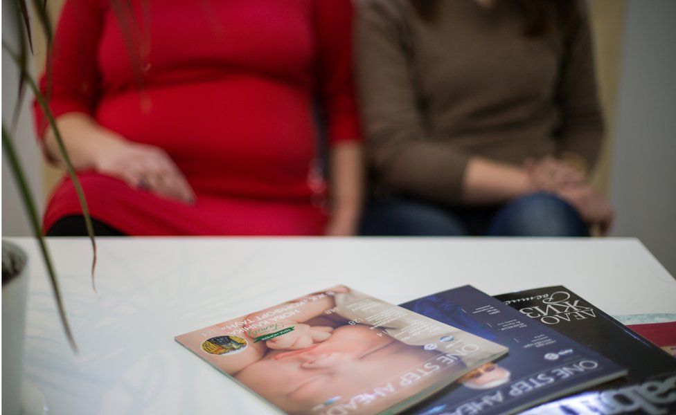 Tetiana and Jana, two surrogates, sit in a waiting room in front of a coffee table with magazines, one of which has a baby's face on the cover