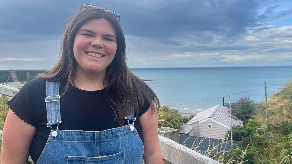 Mared Llywelyn, with long dark hair, smiles at the camera, she is wearing dungarees and the sea is behind her