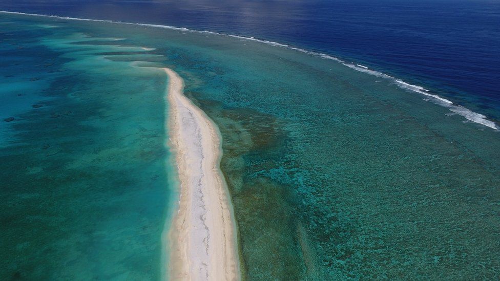 A sand bar stretches forward into a shallow coral reef, with the open ocean beyond
