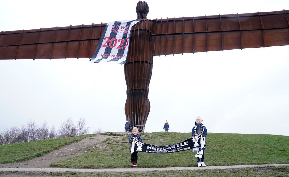 Two boys hold a Newcastle United scarf next to the Angel of the North which has been draped in a giant Newcastle shirt