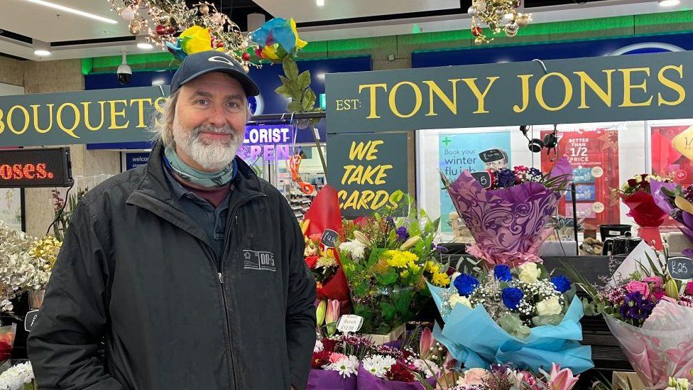 Man with white beard in raincoat standing next to display of bouquets
