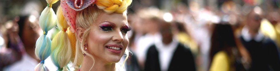 Drag Queen Courtney Act smiles in a rainbow wig and pink makeup