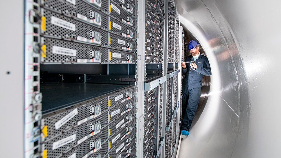 A man in the tight confines of the opened tube works on a computer in a rack of identically sized machines